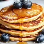 Fluffy stack of protein pancakes recipe topped with fresh blueberries and maple syrup, with syrup drizzling down the sides.